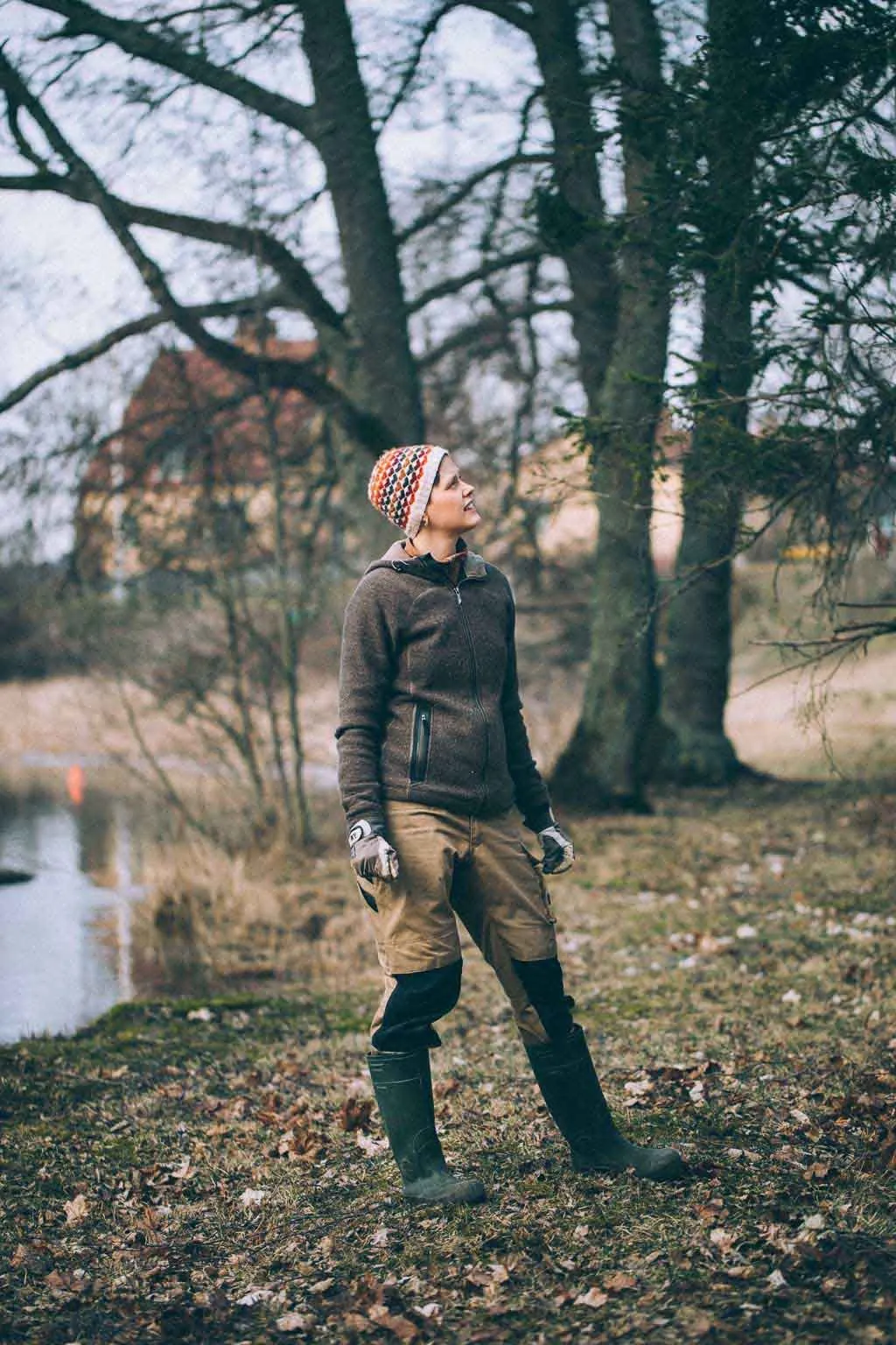 Trädgårdsrådgivare i arbetskläder och mössa vid sjökant omgiven av höstliga träd.